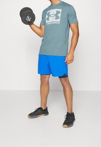 Šedé tričko s krátkými rukávy a velkým logem, modré sportovní kraťasy a černé tenisky s gumovými podrážkami, držící černou činku.