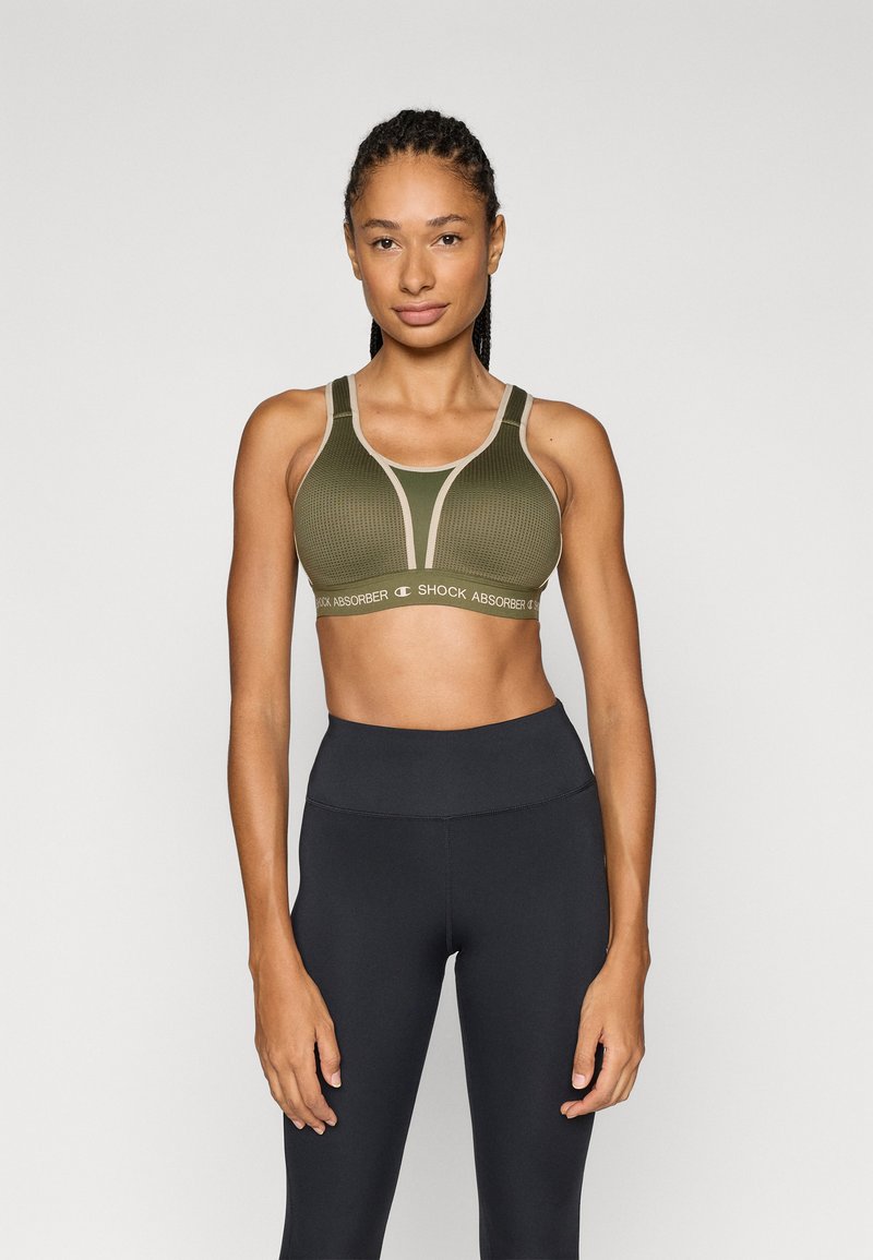 Αθλητικό σουτιέν σε ελιά πράσινο με ραβδωτή υφή, φαρδιές τιράντες και μπεζ λεπτομέρειες, με την ένδειξη "SHOCK ABSORBER" στη λωρίδα κάτω.