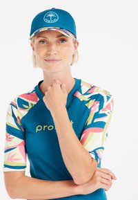 Teal rash guard met kleurrijk tropisch patroon op de mouwen; ronde PROTEST logo pet; gladde stof, korte mouwen, relaxte pasvorm.