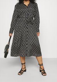 Długa czarna sukienka z białym wzorem w łańcuchy, z paskiem w talii, z zapięciem na guziki, w połączeniu z czarnymi sandałami na cienkich paskach i torebką.