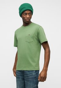 Νεαρός άνδρας που φοράει πράσινο σκουφάκι και ταιριαστό πράσινο κοντομάνικο μπλουζάκι με τσέπη, συνδυασμένο με μπλε τζιν, κοιτάζοντας προς τα πλάγια.