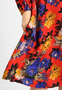 Main tenant l'ourlet d'une robe rouge à gros motif floral jaune, bleu, violet, beige et noir avec manches à poignets élastiques.
