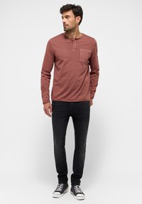 Hosszú ujjú, rozsdabarna henley póló, három gombos pánttal és mellzsebbel, fekete, slim-fit farmernadrággal és sötét tornacipővel párosítva.
