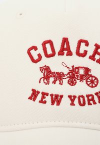 Rode geborduurde tekst "COACH NEW YORK" en een paardenkar op witte stof, waarschijnlijk de voorkant van een pet of hoed.