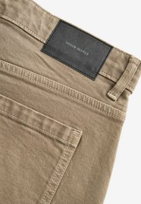 Pantalons en denim beige avec une finition texturée, dotés d'une étiquette en cuir noir marquée "DENIM SUPPLY" et de coutures contrastées.