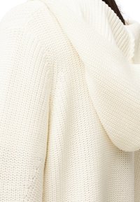 Detailní záběr na krémově zbarvený pletený svetr s kapucí, který má na sobě osoba s tmavohnědými vlasy.