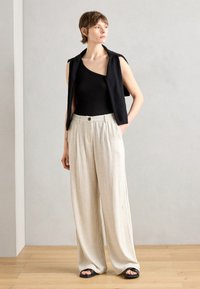 Schwarzes ärmelloses Oberteil, hell gestreifte weite Hose und schwarze Sandalen. Weicher Stoff, lockere Passform und Knopfdetail am Bund.