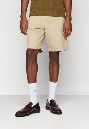 Mand iført beige shorts, hvide ankelsokker med lille logo, mørkebrune læderloafers og en olivengrøn ribstrikket skjorte.