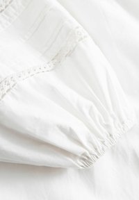 Biała bawełniana tkanina z marszczonym mankietem; ozdobiona delikatną koronką wzdłuż szwu; gładka struktura z lekkim połyskiem.