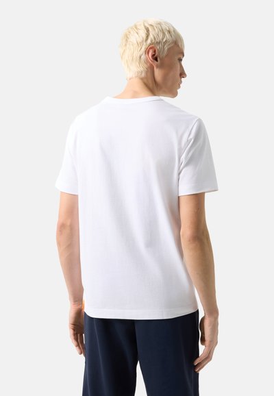 Blond osoba s krátkými vlasy, která má na sobě jednoduché bílé tričko s krátkými rukávy a tmavé kalhoty, stojící zády k prostému pozadí.