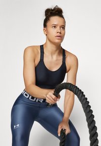 Svart sports-bh med justerbare stropper, kombinert med blå tights med en hvit logo og elastisk midje. Brukeren griper en tykk svart tau.