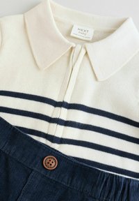 Haut en maille pour bébé couleur crème avec col et rayures bleu marine, associé à un pantalon en velours côtelé bleu marine avec un bouton marron et une taille élastique.