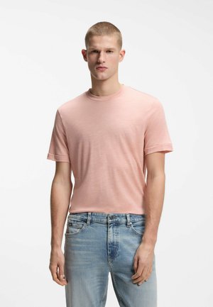 Unge mand iført en lyserød kortærmet t-shirt og lyseblå jeans, der står mod en ren hvid baggrund.