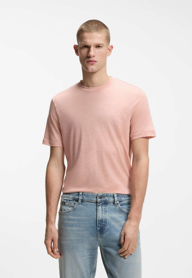 Jeune homme portant un t-shirt rose clair à manches courtes et un jean bleu clair, debout devant un fond blanc uni.