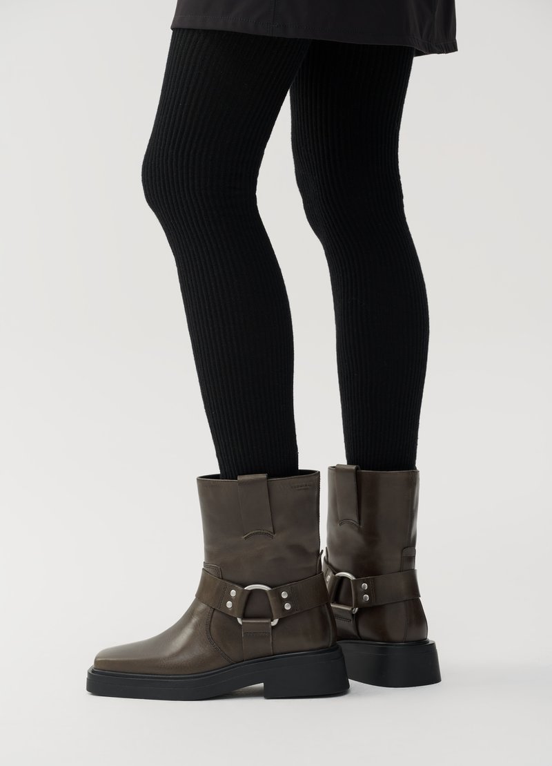 Braune Lederstiefeletten mit abgerundeter Spitze, dicker schwarzer Sohle, seitlichen Schnallenakzenten und glänzender Oberfläche. Getragen mit gerippten schwarzen Leggings.