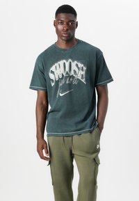 Dark green t-shirt with "Swoosh" graphic and Nike logo; features short sleeves, crew neck, and a faded, textured look. Paired with olive joggers.