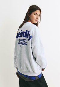 Felpa grigia con vestibilità oversize, polsini e orlo a coste. Presenta un grande testo blu sul retro. Stratificata con magliette blu e scure sotto.