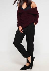 Burgunderroter, gestrickter Pullover mit Carmenausschnitt, kombiniert mit schwarzen, taillierten Hosen; Modell trägt schwarze Ballerinas und präsentiert eine entspannte Passform.