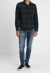 Man i en grön och marinblå rutigt knappad skjorta, blå slim-fit jeans och svarta sneakers med vita sulor, som står mot en vit bakgrund.