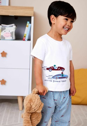 Mosolygó fiatal fiú fehér, versenyautós mintájú pólóban és csíkos nadrágban, aki egy plüss macit tart egy hangulatos hálószobában.