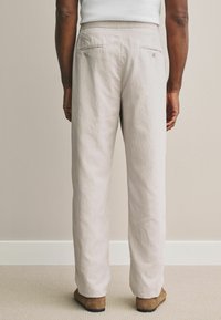 Pantalon beige clair en tissu texturé, avec une coupe décontractée, deux poches arrière avec fermetures à bouton et une jambe droite.
