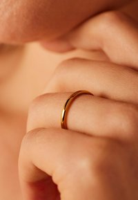 Gouden ring om een vinger, met een gladde, ronde band. Huidtextuur zichtbaar rond de ring, wat het minimalistische ontwerp van de sieraden benadrukt.