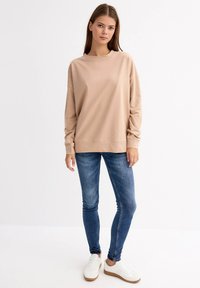 Sudadera de manga larga beige hecha de un tejido suave, combinada con jeans ajustados azules y zapatillas blancas con suela marrón.