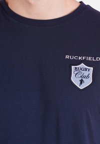 T-shirt en coton bleu marine avec un col rond. Logo en relief représentant un écusson avec l'inscription "RUGBY Club" accompagnée d'une petite icône de joueur. "RUCKFIELD" est imprimé en dessus.