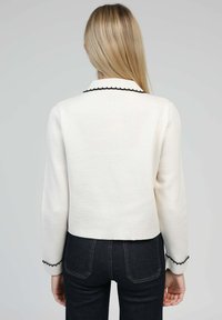 Veste blanche courte avec des bordures festonnées noires au col et aux poignets. Le tissu a une finition texturée. Associée à un pantalon en denim foncé.