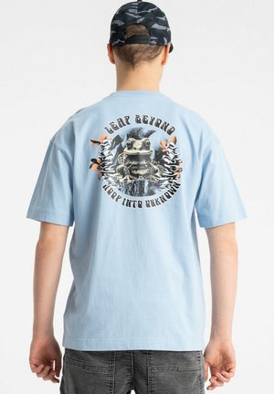 Osoba nosząca jasnoniebieski t-shirt z grafiką żaby i napisem "Leap beyond, hop into unknown" na plecach, w połączeniu z czarną czapką moro i szarymi dżinsami.