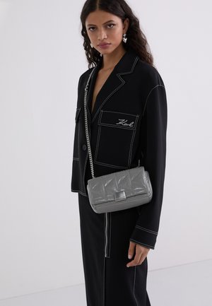 Blazer nero con cuciture bianche e tasche frontali, abbinato a una borsa grigia trapuntata con tracolla in catena argentata.