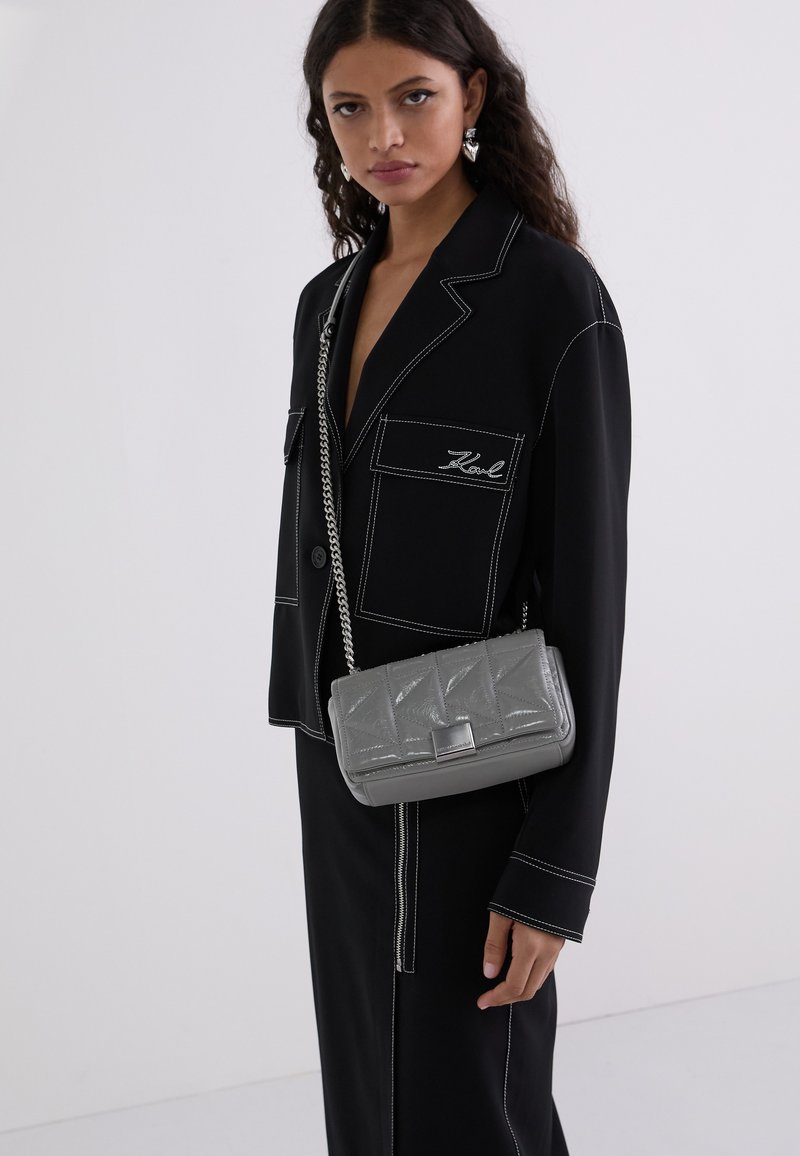 Blazer nero con cuciture bianche e tasche frontali, abbinato a una borsa grigia trapuntata con tracolla in catena argentata.