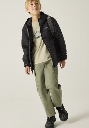 Blond jongetje draagt een zwarte pufferjas, beige T-shirt met print, groene cargobroeken en zwarte sneakers, lopend met een rugzak op een beige achtergrond.
