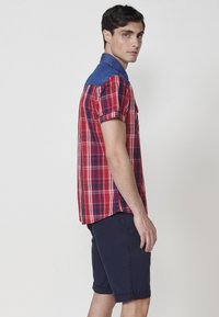 Kurzärmliges kariertes Hemd in Rot, Blau und Weiß; Denim-Akzente an den Schultern. Kombiniert mit marineblauen Shorts mit umgeschlagenen Säumen.