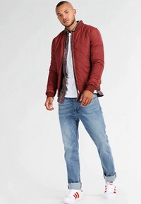 Chaqueta bomber de acolchado rojo con puños acanalados, camiseta blanca, camisa de cuadros debajo, jeans azul claro y zapatillas blancas con detalles rojos.