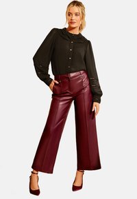 Vrouw in een zwarte blouse met lange mouwen en hartknopen, gecombineerd met bordeauxrode leren wijde broek, staande met een hand in de zak.