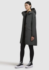 Manteau noir résistant à l'eau de couleur gris foncé avec une capuche, doté de boutons-pression, de poches latérales et d'une coupe droite. Associé à des leggings noirs et des baskets.