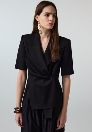 Frau mit langen braunen Haaren trägt schwarzen kurzärmligen Blazer, große goldene Hängeohrringe und braune Armbänder, leicht nach rechts blickend vor grauem Hintergrund.