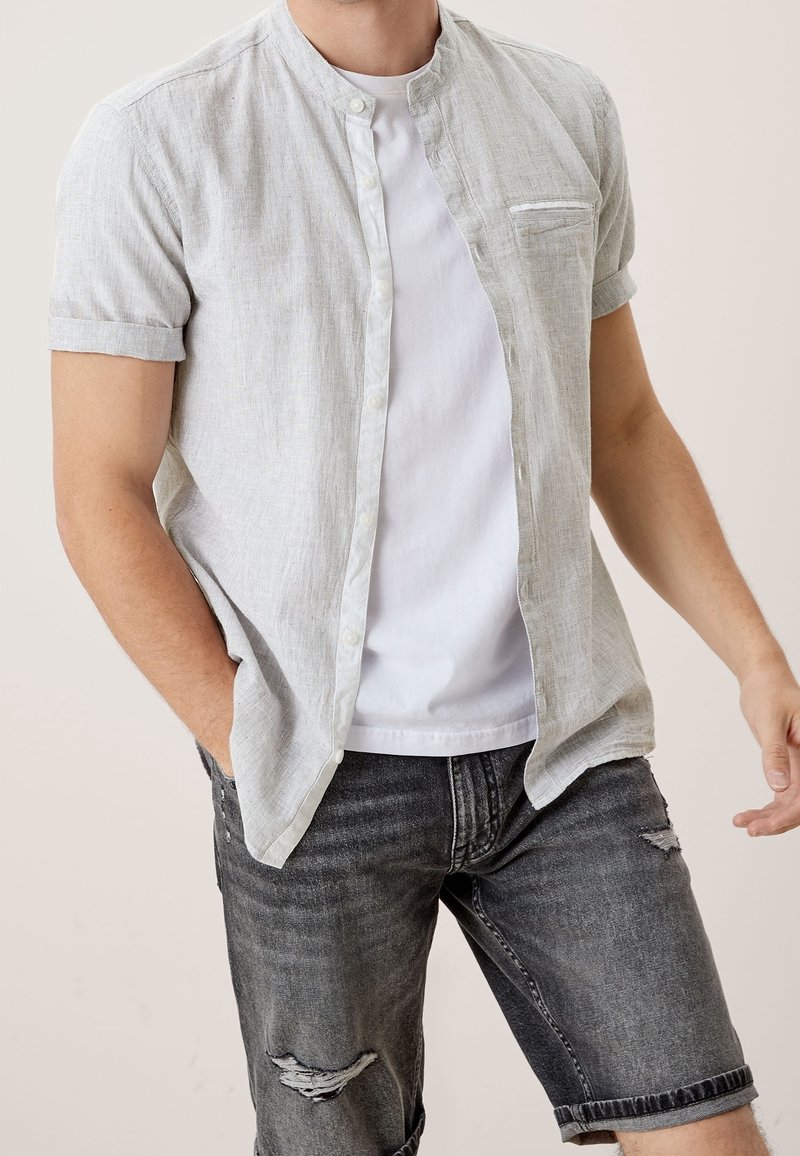 Homme portant une chemise à manches courtes gris clair boutonnée sur un t-shirt blanc et un short en jean gris foncé déchiré, posant avec une main dans la poche.