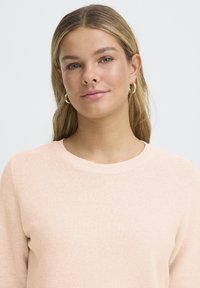 Gaiši rozā adīts džemperis ar apaļu izgriezumu un ribas aprocēm. Mīksta tekstūra un slaids piegriezums, piemērots kā kārtas slānis vai ikdienas valkāšanai.