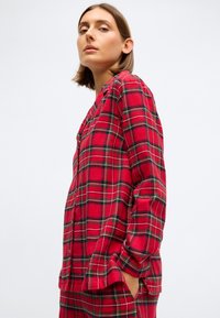 Rød ternet flannel skjorte med lange ærmer, knapfront og en afslappet pasform. Mønsteret inkluderer sorte og grønne detaljer.