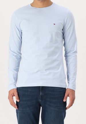 Hellblaues Langarm-T-Shirt aus glattem Stoff mit rundem Ausschnitt und kleinem ikonischem Logo auf der linken Brust. Kombiniert mit dunklen Jeans.