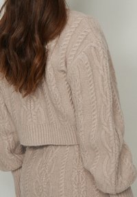 Cardigan en maille torsadée de couleur beige doux. Présente une coupe courte, un ourlet large côtelé et un fil texturé avec des motifs en relief le long des manches.