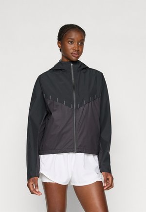 Zwarte lichtgewicht jas met capuchon, voorzien van een diagonaal ontwerp, ritssluiting en subtiele reflecterende accenten. Draagbare over witte shorts.