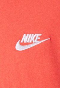 Koralové bavlněné tričko s texturovaným bílým vyšívaným logem Nike na přední straně, na hladkém, rovnoměrném látkovém pozadí.