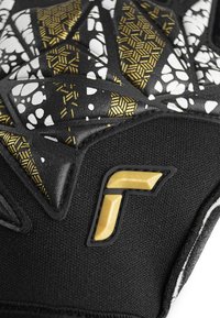 Guante deportivo negro con agarre texturizado, que presenta patrones geométricos blancos y dorados. Detalle destacado de logo dorado y textura de tela contrastante.