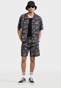 Black short-sleeve shirt and shorts set with white graphic patterns. Accompanied by a black bucket hat and sunglasses, black sneakers with white accents.