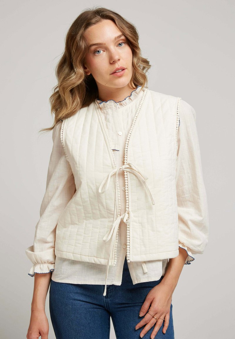 Femme aux cheveux ondulés portant un gilet matelassé crème noué devant sur une blouse légère à volants et un jean bleu, devant un fond uni.