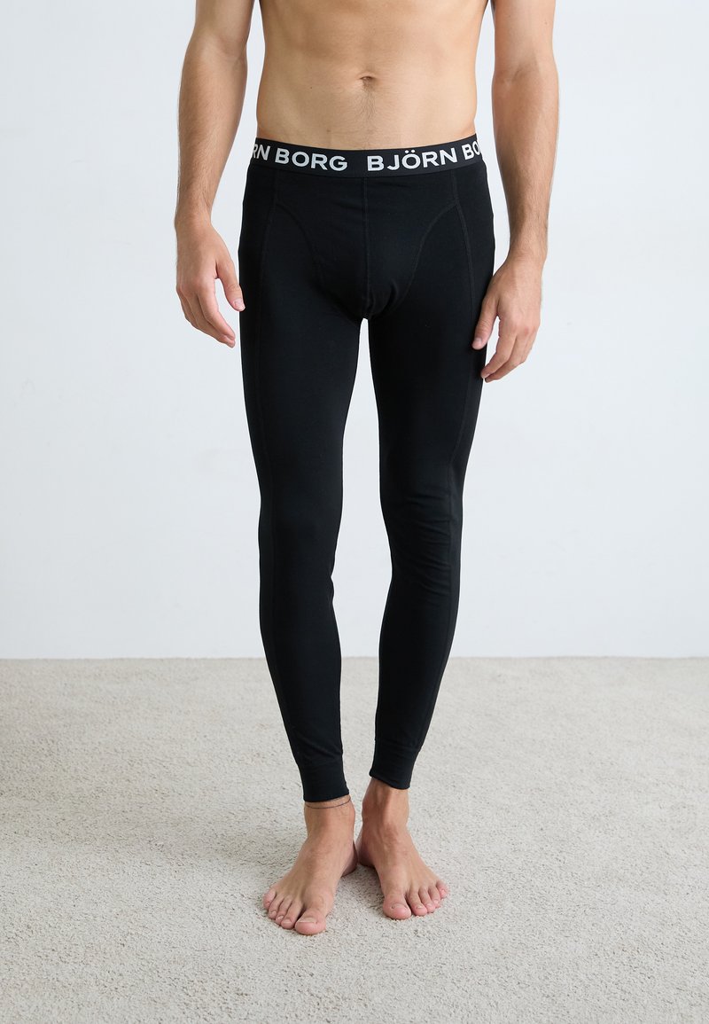 Leggings em tecido elástico preto com um ajuste justo, apresentando uma bainha de logo branco. Textura suave e design afunilado que se estende até os tornozelos.