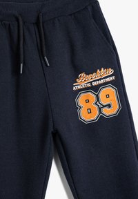 Marineblå svedbukser lavet af blødt stof, udstyret med elastisk talje og snøre samt farverig grafik med "Brooklyn Athletic Department" og nummer 89.
